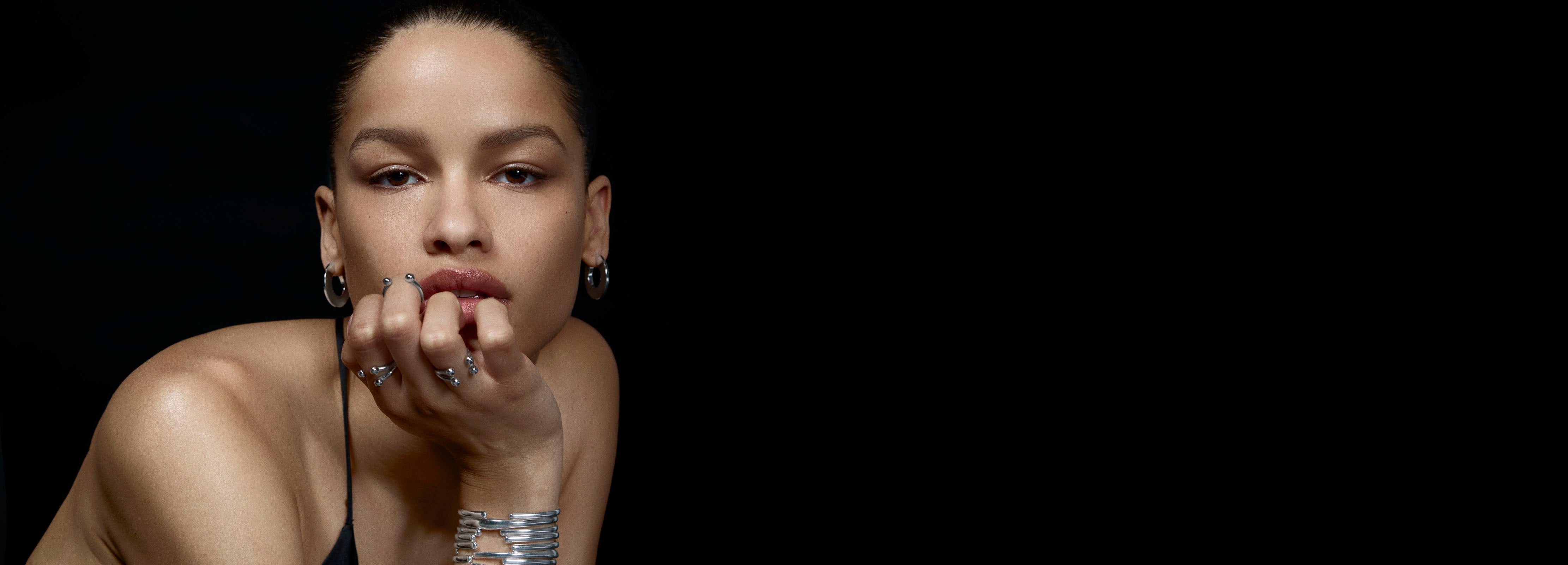 Woman with hand on chin against a black background wearing silver bracelets and rings